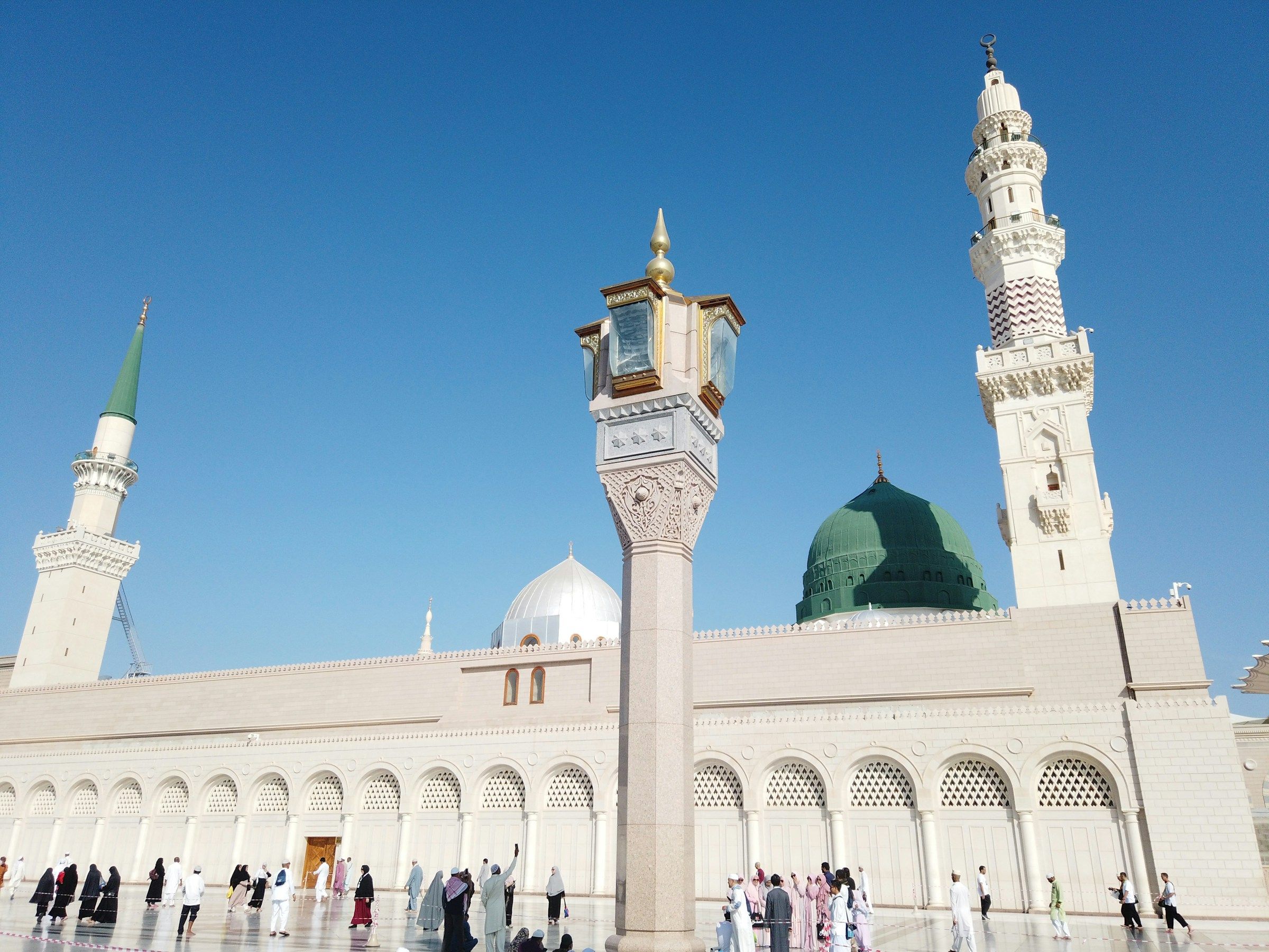 The Green Dome and minarets of Al-Masjid an-Nabawi under a clear sky, with pilgrims in the courtyard